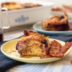 A slice of lemon blueberry croissant bake on a plate with a side of bacon.