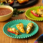 A table with two plates of stuffed mini peppers and a bowl of salsa.